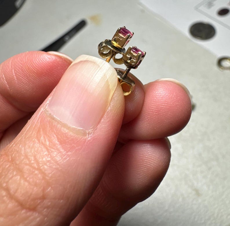 May include: A pair of small stud earrings featuring round, faceted pink-red gemstones set in gold-toned metal prongs. The earrings have a post-back design with visible metal butterfly clutch backings attached to the posts behind the stone settings.