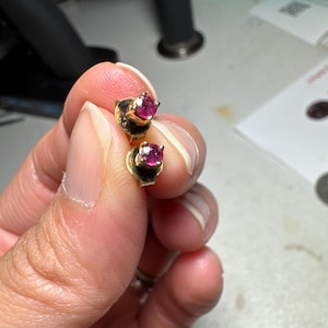 May include: A pair of gold stud earrings, each featuring a single round-cut, deep pinkish-red gemstone held in a four-prong setting. The earrings are small in size and have a polished gold finish, showcasing a classic design with a vibrant center stone.