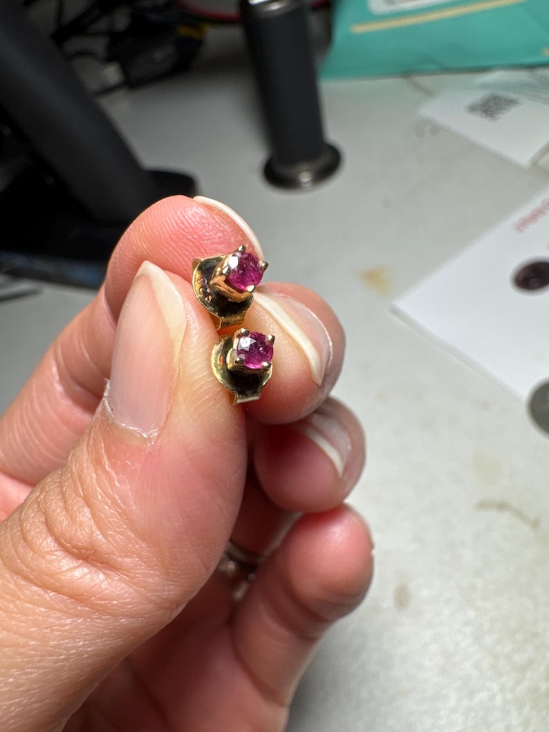 May include: A pair of gold stud earrings, each featuring a single round-cut, deep pinkish-red gemstone held in a four-prong setting. The earrings are small in size and have a polished gold finish, showcasing a classic design with a vibrant centre stone.