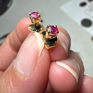 May include: A pair of stud earrings featuring round, faceted, vibrant pink gemstones set in gold-toned metal prongs. Each earring includes a matching gold-toned metal backing, showcasing a classic design with a polished finish and secure setting.