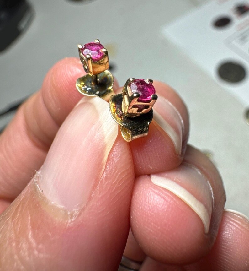 May include: A pair of stud earrings featuring round, faceted, vibrant pink gemstones set in gold-toned metal prongs. Each earring includes a matching gold-toned metal backing, showcasing a classic design with a polished finish and secure setting.