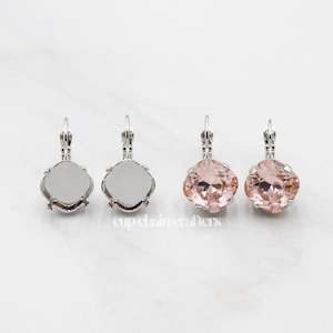 May include: Four earrings are displayed. Two are silver-toned with a square setting, and two are a light pink with a faceted, cushion-cut design. The earrings have a lever-back closure. The background is a white marble surface.