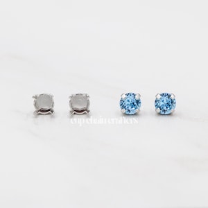 May include: Four stud earrings. Two are silver-colored settings without stones. The other two are silver-colored settings with round, light blue gemstones. The earrings are displayed on a white surface.