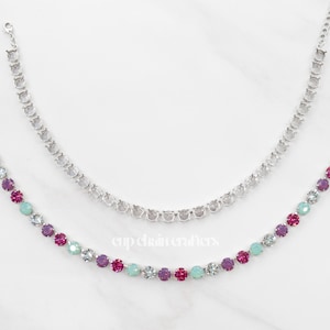 May include: Two necklaces with silver chains. One is adorned with clear crystals, the other with a row of colorful crystals in shades of purple, pink, and teal. The necklaces are displayed on a white surface.