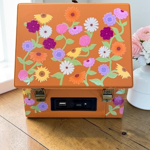 May include: An orange rectangular box with a hinged lid featuring a colorful floral pattern of yellow, pink, purple, and white flowers with green stems. The front panel includes a black recessed port area with a USB and a USB-C connection.