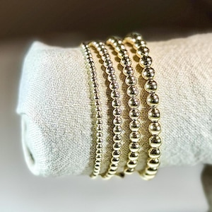 May include: Five gold beaded bracelets stacked on top of each other. The bracelets are made of small, round beads.