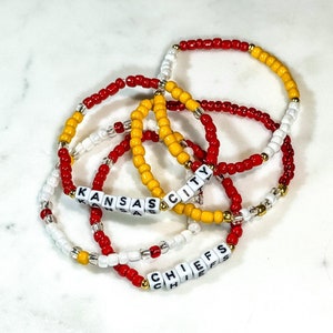 May include: Three beaded bracelets with the words "KANSAS CITY" and "CHIEFS" spelled out in white letter beads. The bracelets are red, white, and yellow.