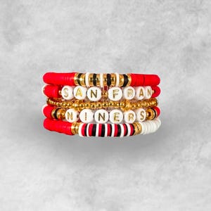 May include: Stack of beaded bracelets in red, white, black, and gold. The bracelets spell out "SAN FRAN" and "NINERS" in gold letter beads. The bracelets are stacked together, creating a layered look.