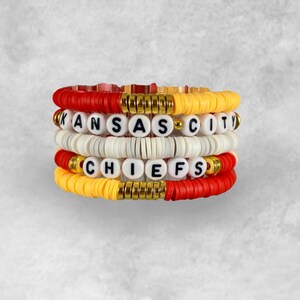 May include: A set of four beaded bracelets with the words "KANSAS CITY CHIEFS" spelled out in black letters on white beads. The bracelets are made with red, yellow, and gold beads.