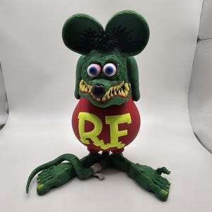 Può includere: Una figura di ratto verde e caricaturale con grandi orecchie, una pancia rossa con le lettere "RF" in giallo e lunghi piedi verdi artigliati. Il ratto ha un'espressione minacciosa con denti affilati e occhi sporgenti.