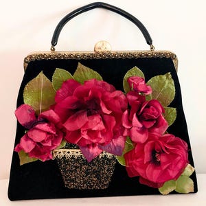 May include: Black velvet handbag with a decorative floral arrangement. The bag features a black handle and a gold-toned clasp. The floral arrangement includes red roses, green leaves, and a black beaded basket.