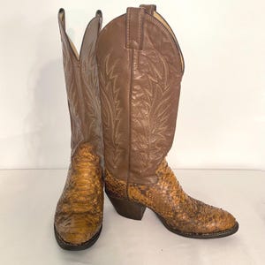 May include: A pair of tall cowboy boots featuring a brown leather shaft with decorative stitching and a textured, snakeskin-patterned leather foot. The boots have a pointed toe, a stacked heel, and pull tabs at the top of the shaft for easy wear.