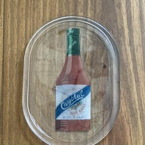 May include: Clear, oval-shaped coaster featuring a graphic of a Crystal Hot Sauce bottle. The bottle is red with a green cap and a blue and white label. The coaster is on a wooden surface.