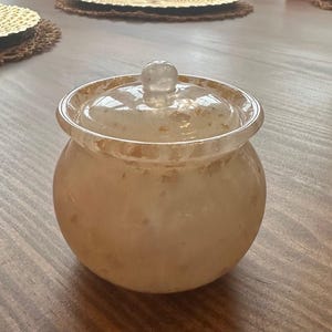 May include: A white ceramic jar with a gold lid. The jar has a round shape and a smooth, glossy finish. The lid has a small knob on top.