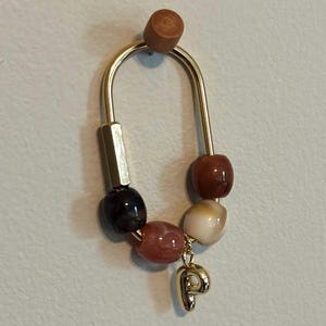 May include: A gold-colored earring with a curved design, featuring a series of beads in shades of brown, red, and cream. A small gold charm shaped like the letter "P" dangles from the earring.