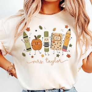 May include: An ivory t-shirt featuring a graphic of a green crayon, an orange pumpkin, a green glue stick, a spiral notebook, and a yellow pencil, all with smiling faces. The text "mrs. taylor" is printed in orange cursive below the characters.