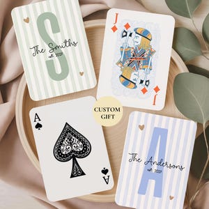 May include: Four playing cards arranged on a wooden tray. One card features a large green letter S with the text The Smiths est. 2027. Another shows a blue letter A with The Andersons est. 2027. A third is an Ace of Spades, and the fourth is a Jack of Diamonds.