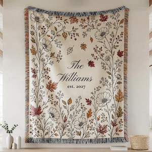May include: A cream-colored woven throw blanket featuring a botanical pattern of black-outlined flowers and autumn-toned leaves. The center displays the text The Williams est. 2027 in a black serif font. The blanket has a fringed edge on all sides.