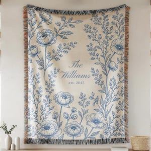 May include: A woven throw blanket featuring a cream-colored background with blue floral illustrations. The center displays the text The Williams est. 2027. The blanket has fringed edges with multicolored threads along the top and bottom borders.