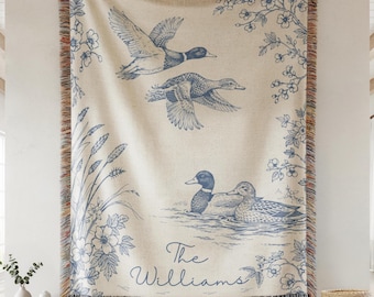 May include: A woven throw blanket featuring a cream background with blue illustrations of ducks in flight and swimming on water. The design includes floral accents, reeds, and the text The Williams in a script font, finished with fringed edges.