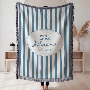 May include: A blue and white striped throw blanket with a fringed edge. The blanket features an oval design with the text "The Johnsons est. 2026" in a script font. The blanket is made of a woven material.