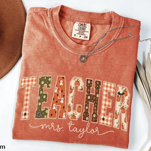 May include: A rust-colored short-sleeve t-shirt with a crew neckline. The front features the word TEACHER in block letters with various autumn-themed patterns, including pumpkins, ghosts, and plaid. Below, the text mrs. taylor is printed in white.