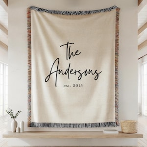 May include: Cream-colored woven throw blanket with black fringe and the text "The Andersons est. 2015" in a script font. The blanket has a decorative border with red, blue, and tan accents. A personalized home decor item.
