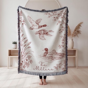 May include: Cream-colored woven throw blanket with a burgundy duck illustration. The design features ducks in flight and swimming, with floral and plant accents. The text "The Millers" is at the bottom. The blanket has a fringed edge.