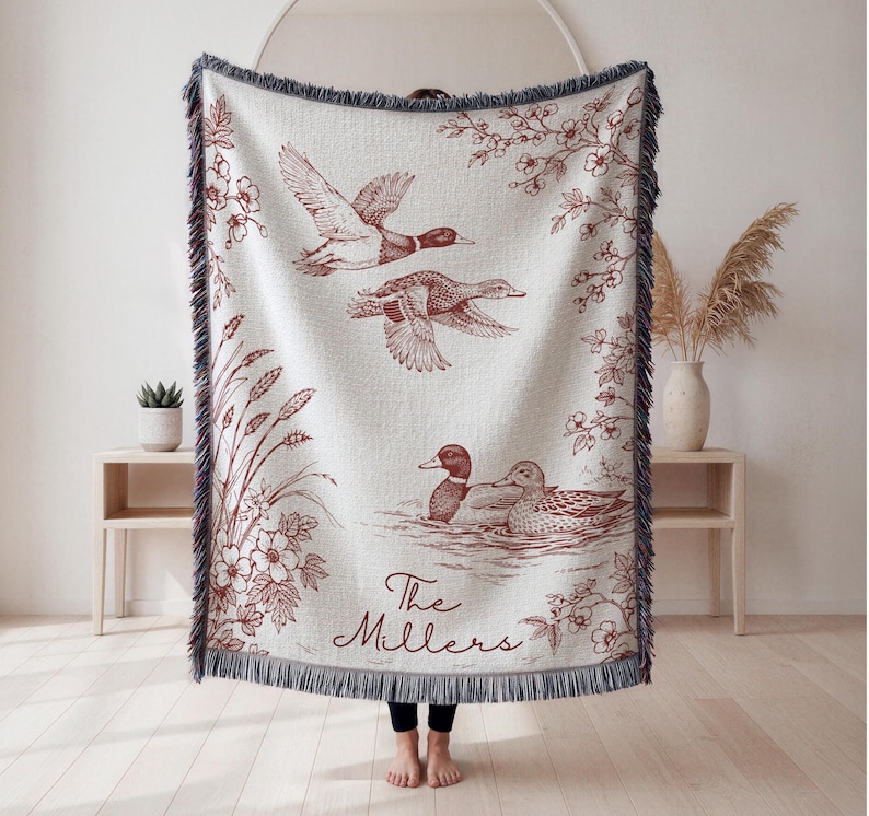 May include: Cream-colored woven throw blanket with a burgundy duck illustration. The design features ducks in flight and swimming, with floral and plant accents. The text "The Millers" is at the bottom. The blanket has a fringed edge.