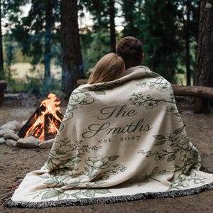 May include: A beige blanket with a floral design and the text "The Smiths est. 2027" is draped over two people. The blanket has a fringed edge and is set in front of a campfire in a forest setting.