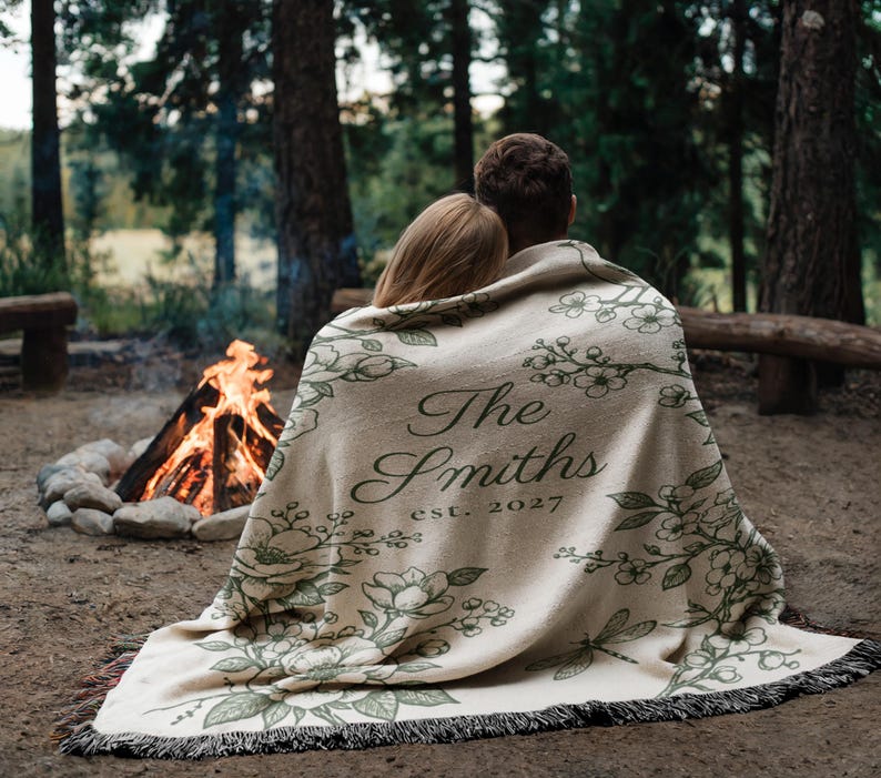 May include: A beige blanket with a floral design and the text "The Smiths est. 2027" is draped over two people. The blanket has a fringed edge and is set in front of a campfire in a forest setting.