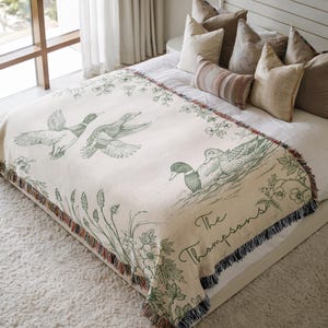 May include: Cream-colored throw blanket featuring a duck illustration in green. The blanket has a fringed edge with a floral and plant design. The text "The Thompsons" is also visible on the blanket.
