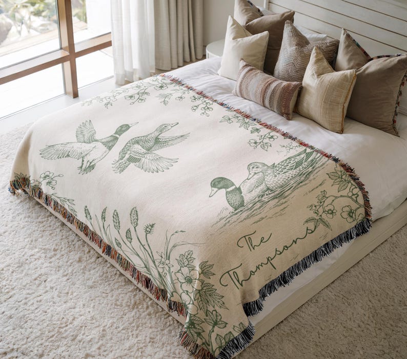 May include: Cream-colored throw blanket featuring a duck illustration in green. The blanket has a fringed edge with a floral and plant design. The text "The Thompsons" is also visible on the blanket.