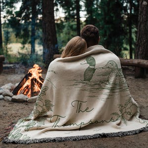 May include: A beige throw blanket with a duck illustration and the text "The Thompsons" in green. The blanket has a fringed edge and is draped over two people sitting near a campfire in a forest setting.
