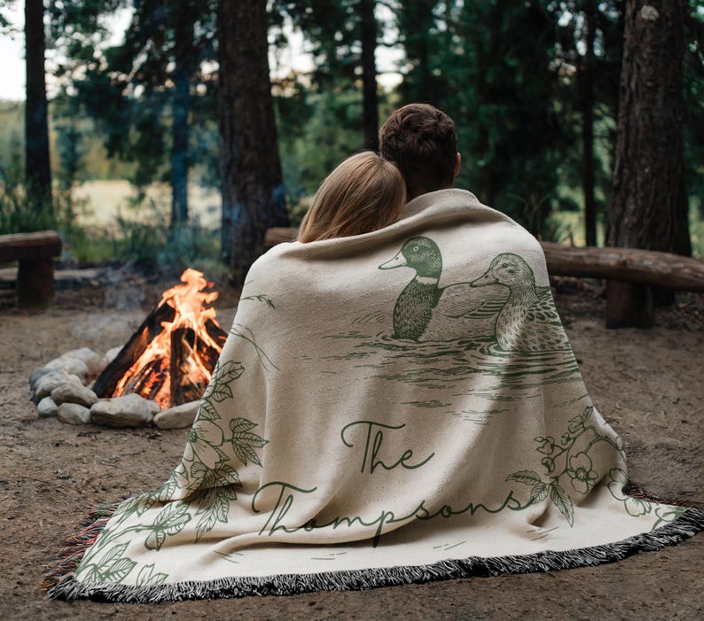 May include: A beige throw blanket with a duck illustration and the text "The Thompsons" in green. The blanket has a fringed edge and is draped over two people sitting near a campfire in a forest setting.