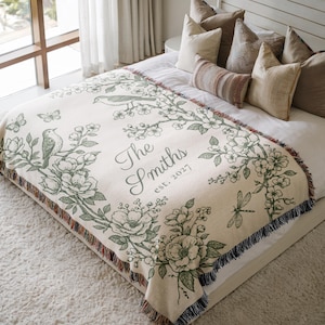May include: Cream-colored throw blanket with a floral and bird design in green. The blanket features the text "The Smiths est. 2027". The blanket has a fringed edge and is displayed on a bed with pillows.