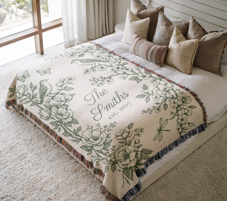 May include: Cream-colored throw blanket with a floral and bird design in green. The blanket features the text "The Smiths est. 2027". The blanket has a fringed edge and is displayed on a bed with pillows.