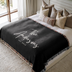 May include: A black woven throw blanket with white script text that reads "The Andersons est. 2015." The blanket has a fringed edge with a multicolored pattern and is draped over a bed with pillows.