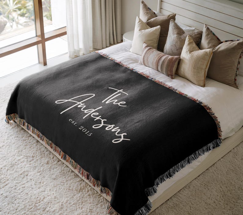 May include: A black woven throw blanket with white script text that reads "The Andersons est. 2015." The blanket has a fringed edge with a multicolored pattern and is draped over a bed with pillows.