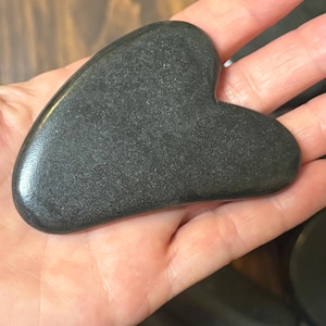 Peut inclure: Un outil de gua sha lisse en forme de cœur, fabriqué en pierre gris anthracite avec une surface polie et réfléchissante. L'objet présente des bords arrondis et une forme ergonomique incurvée, affichant une texture gris foncé uniforme.