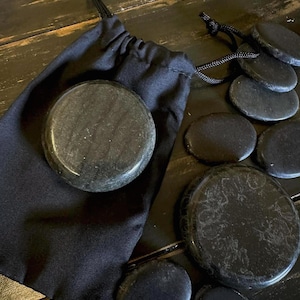 May include: A black drawstring bag with a large, round, dark stone inside, surrounded by several smaller, oval-shaped stones. The stones have a polished, reflective surface. The background is a dark wooden surface.