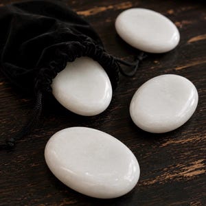May include: Four smooth, oval-shaped white stones with a polished finish. Three of the stones are arranged separately, while one stone is partially emerging from a black drawstring pouch made of a soft, velvet-like fabric with a black cord.