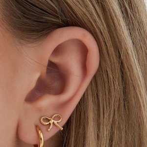 Könnte beinhalten: Nahaufnahme eines Ohrs mit goldenen Ohrringen. Die Ohrringe umfassen einen kleinen Schleifenstecker und einen Creolen-Ohrring. Der Schmuck ist goldfarben, und der Hintergrund sind die Haare der Person.