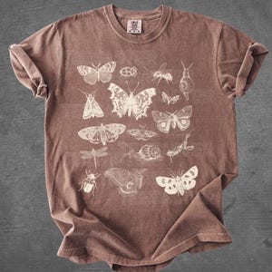May include: Espresso-colored t-shirt featuring a collection of insect illustrations in white. The design includes various butterflies, moths, and other insects. The shirt has a crew neck and short sleeves. The word "ESPRESSO" is printed at the bottom.