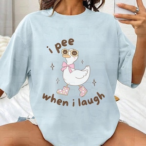 May include: Light blue t-shirt with a graphic of a white goose wearing sunglasses, a pink bow, and boots. The text "i pee when i laugh" is above and below the goose in brown lettering. The shirt is a casual style.