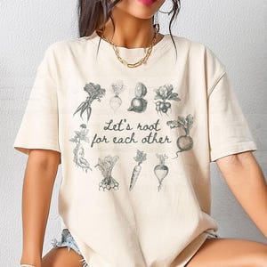 May include: Ivory-colored t-shirt with a botanical illustration of various root vegetables. The text "Let's root for each other" is printed in a script font. The shirt is a relaxed fit.