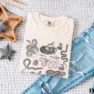 May include: An ivory-coloured t-shirt with a black and white snake illustration, folded on a white quilted surface. Blue denim jeans are next to the shirt. Decorative star-shaped objects are also present. The word 'IVORY' is visible.