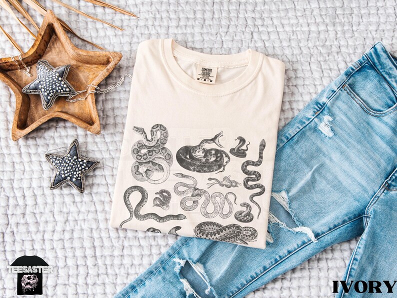 May include: An ivory-coloured t-shirt with a black and white snake illustration, folded on a white quilted surface. Blue denim jeans are next to the shirt. Decorative star-shaped objects are also present. The word 'IVORY' is visible.