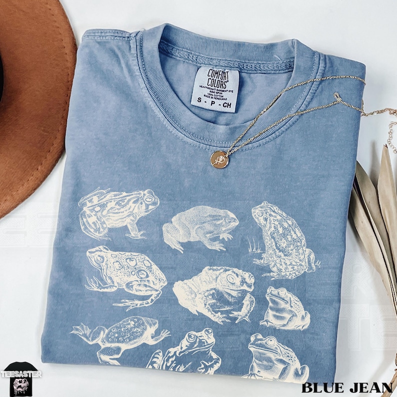 Pu&ograve; includere: T-shirt blu jeans con illustrazioni di rane in varie pose. La maglietta ha un girocollo e maniche corte. Una collana d'oro con un ciondolo &egrave; drappeggiata sulla maglietta. Il testo "BLUE JEAN" &egrave; in basso.