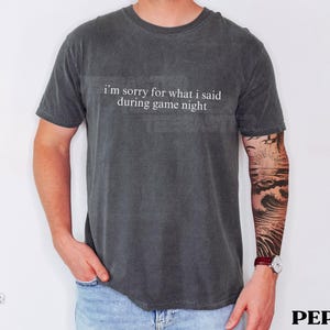 May include: Dark gray t-shirt with the text "I'm sorry for what I said during game night" in white. The shirt is a crew neck style and is worn by a person. The person is wearing blue jeans and has a tattoo on their arm.